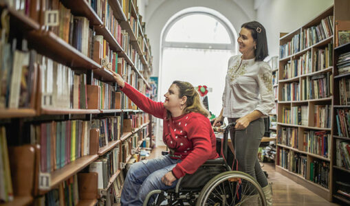 Frau in Rollstuhl und weitere Frau in Bibliothek